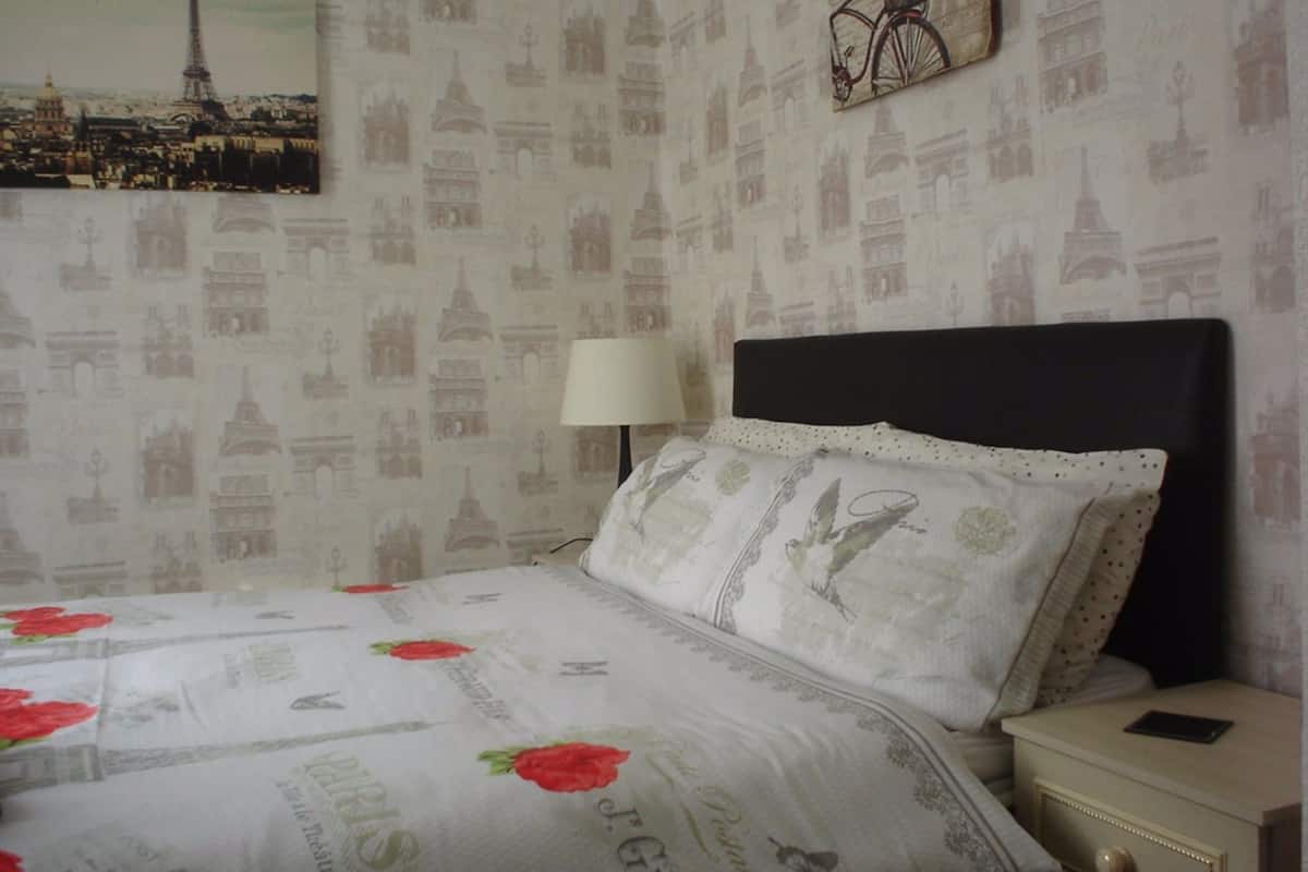 A bedroom with a bed, a nightstand, a lamp, and a wall with Paris-themed wallpaper.