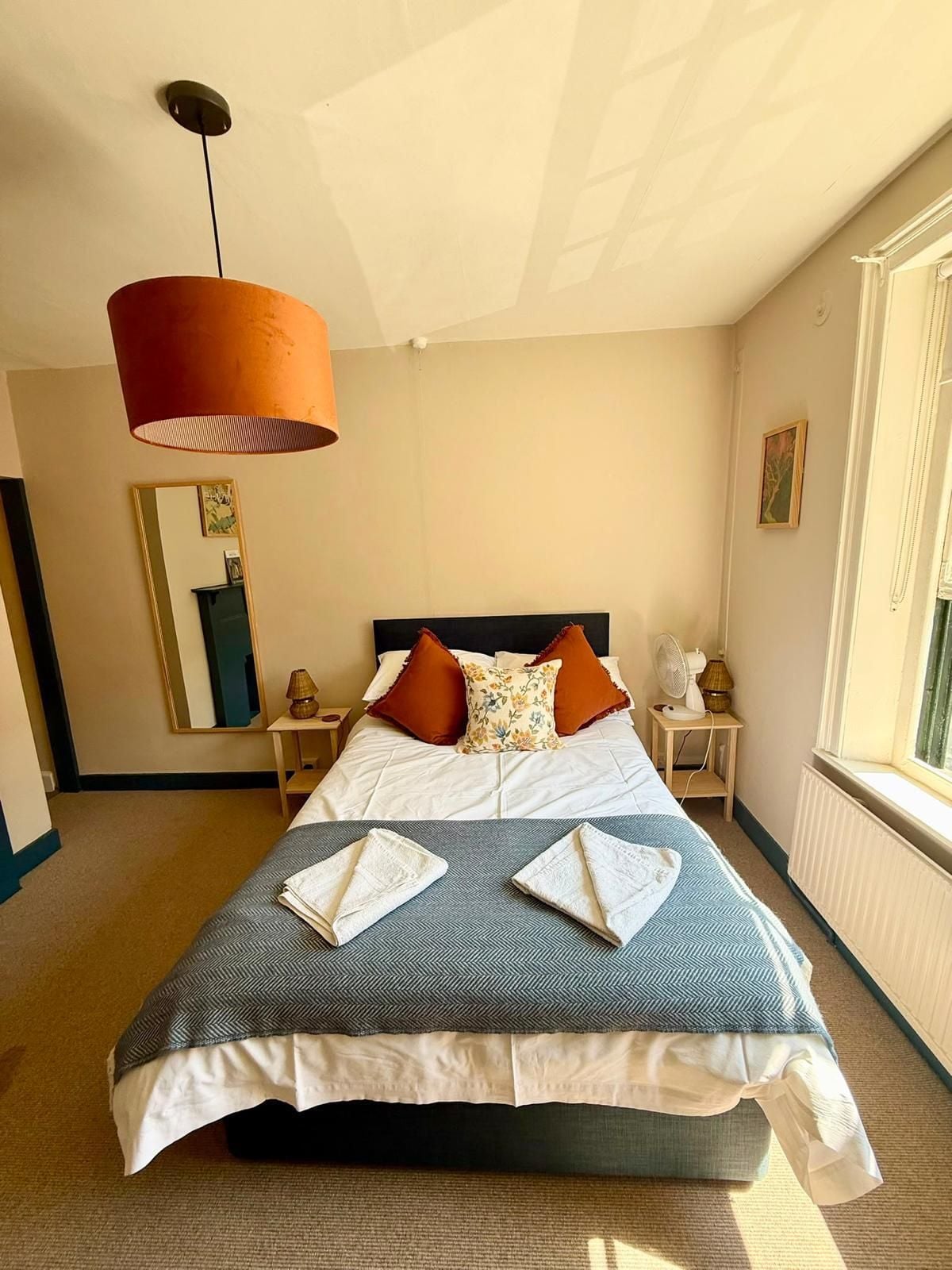A hotel room with a large bed, a standing heater, a bedside table with a lamp, and a window with a view.