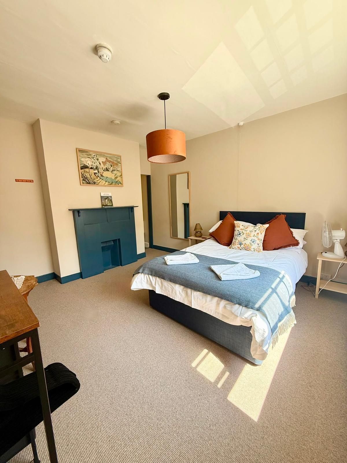 A hotel room with a bed, a desk, a mirror, a fireplace, and a ceiling light fixture.