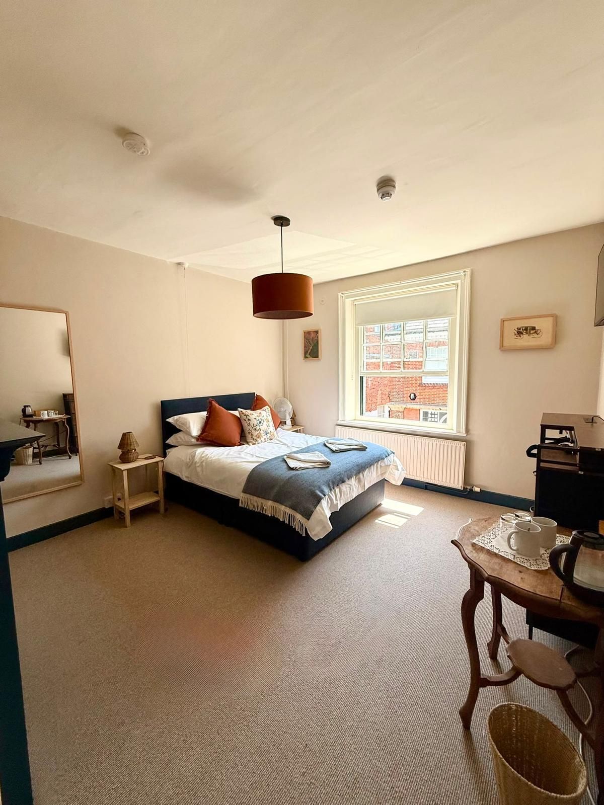 Un dormitorio con cama, una mesa con tazas y una tetera, una ventana con vistas, y una lámpara colgante.