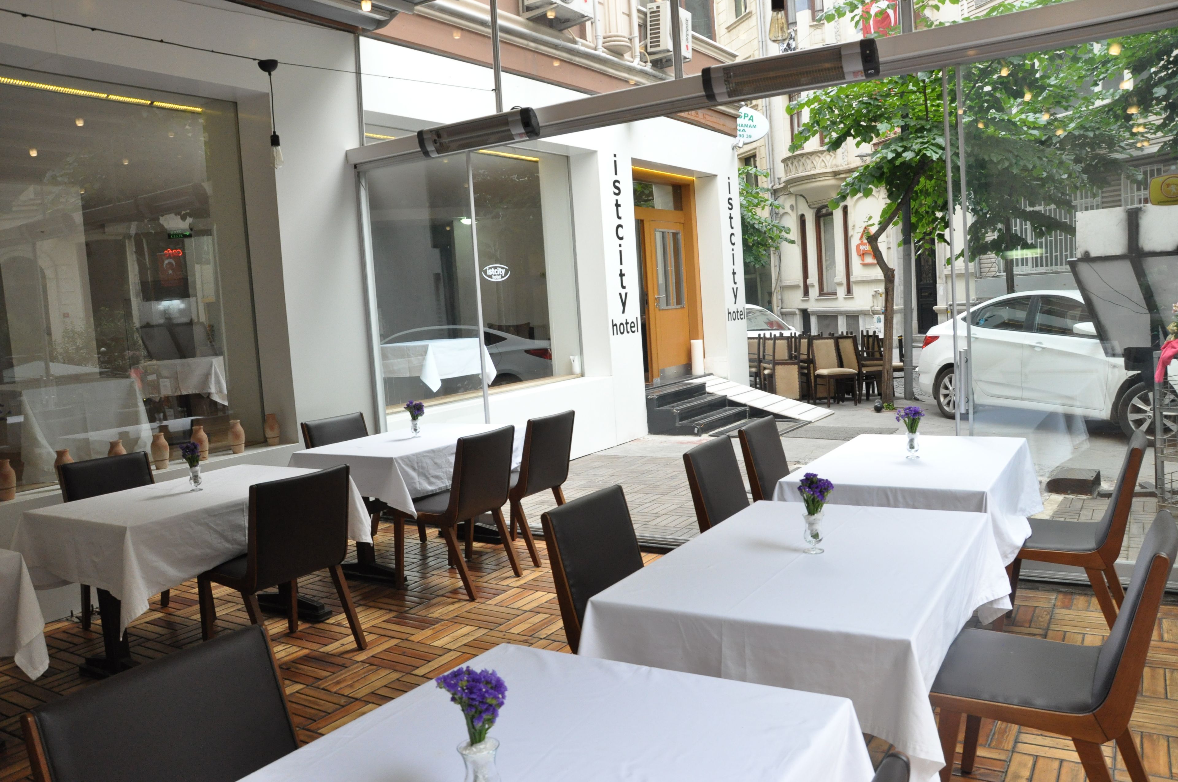 An outdoor seating area with tables and chairs, a building entrance, and a parked car.