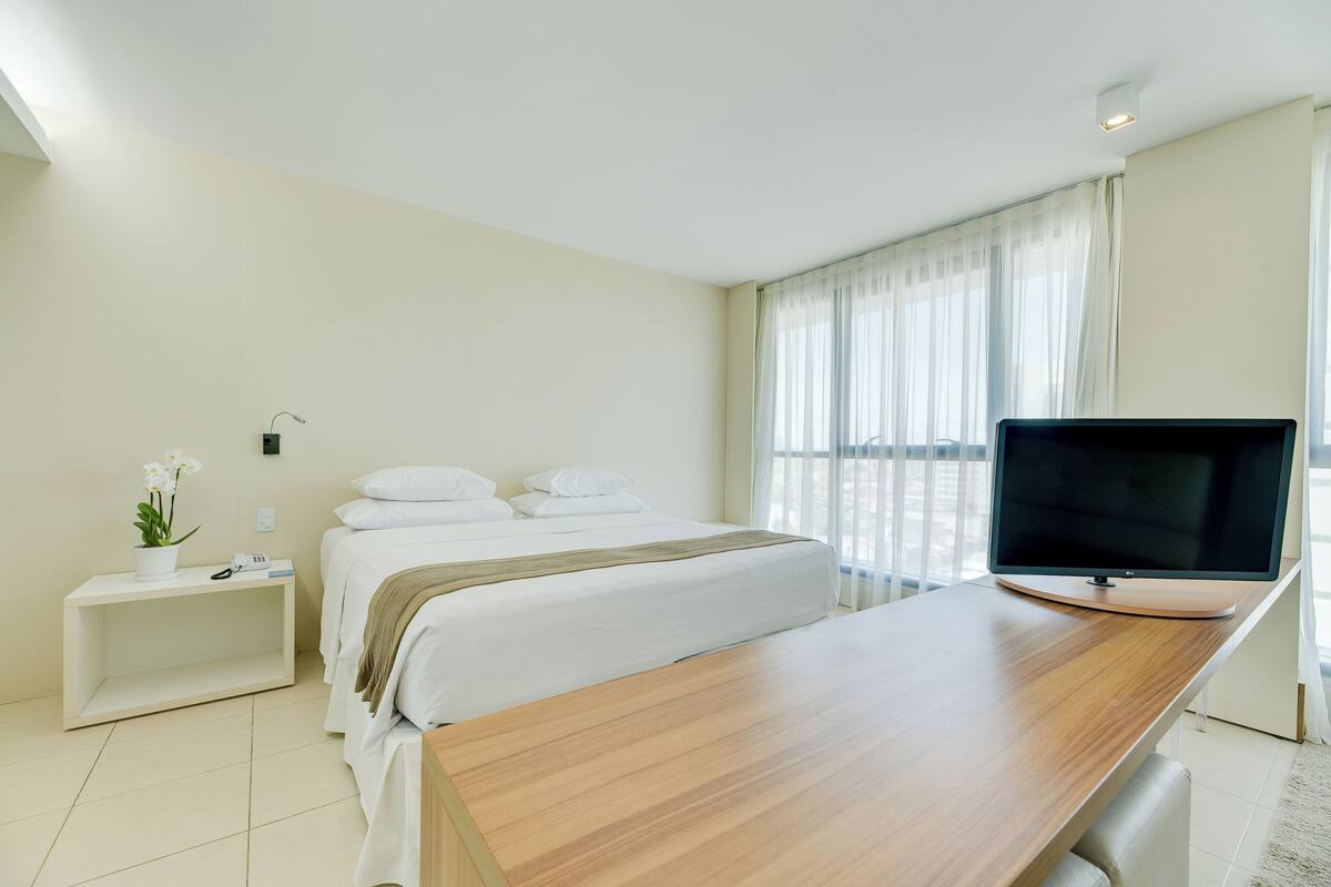 A hotel room with a bed, a bedside table with a plant, a TV on a wooden desk, and a window with curtains.