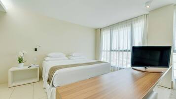 Habitación de hotel con una cama, una mesita de noche con una planta, un televisor sobre un escritorio de madera y una ventana con cortinas.