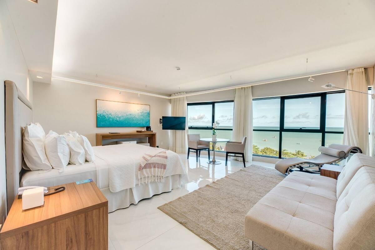 A modern hotel room with a large bed, a sofa, a desk, and a TV. The room has a large window with a view of the ocean.