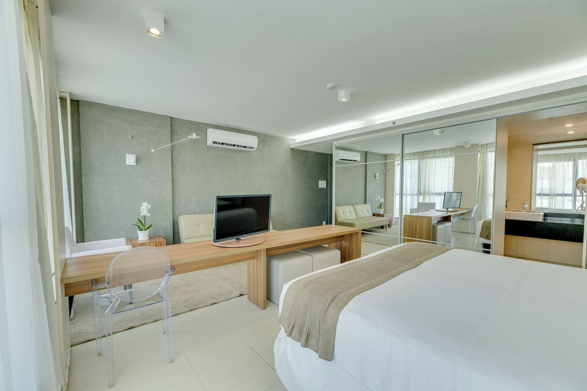 A modern hotel room with a large bed, a wooden desk, a television, and a bathroom visible through a glass partition.