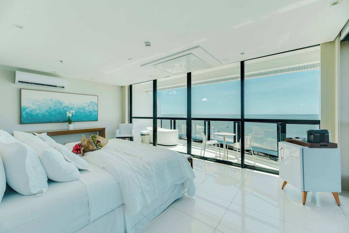 A modern bedroom with a large bed, a desk, and a view of the sea.