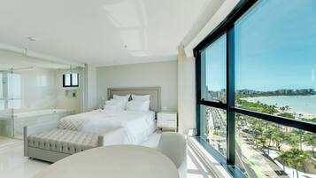 Un dormitorio moderno con una cama grande, ducha de vidrio y vistas a la playa y al paisaje urbano.