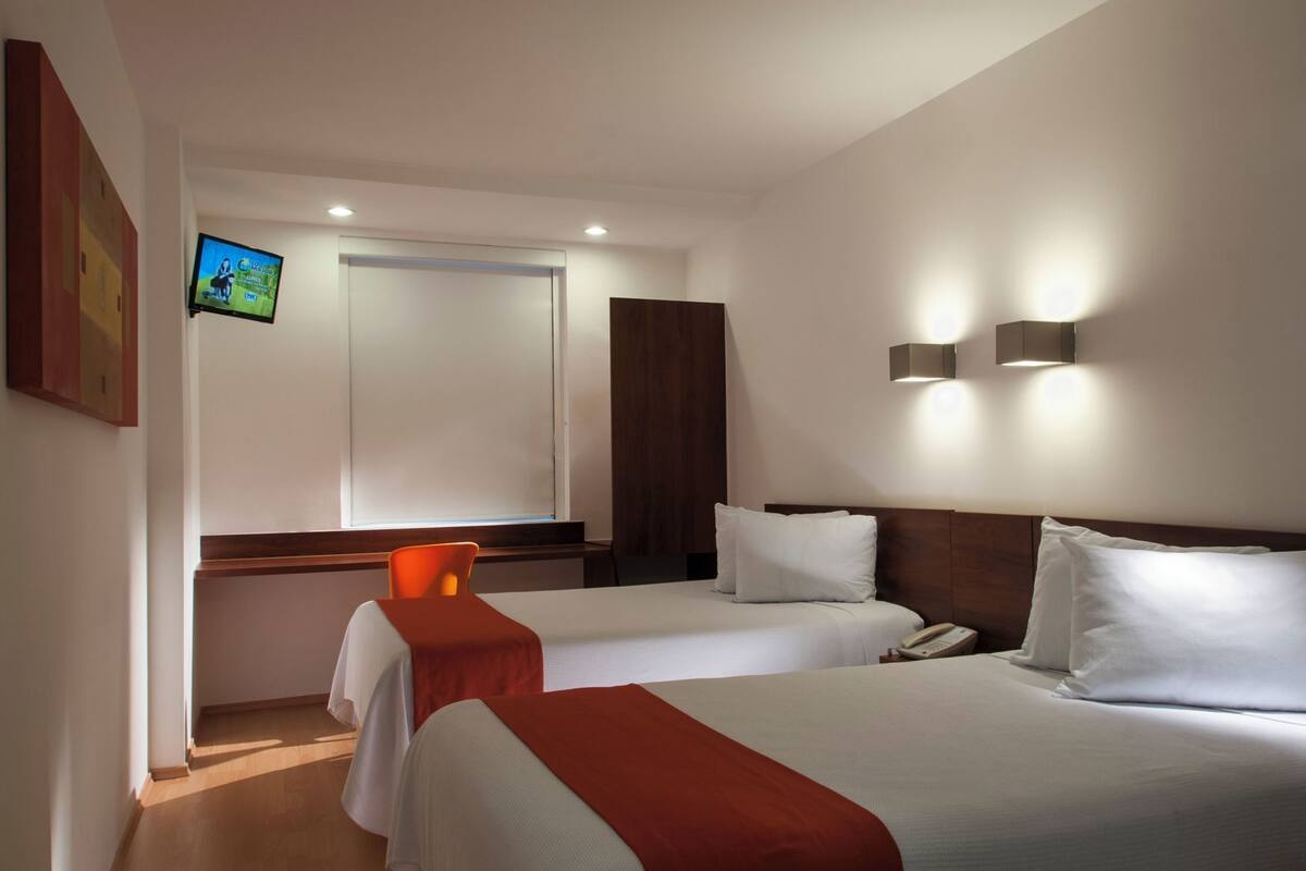 Habitación de hotel con dos camas, una mesita, una silla, un televisor y una lámpara fijada a la pared.