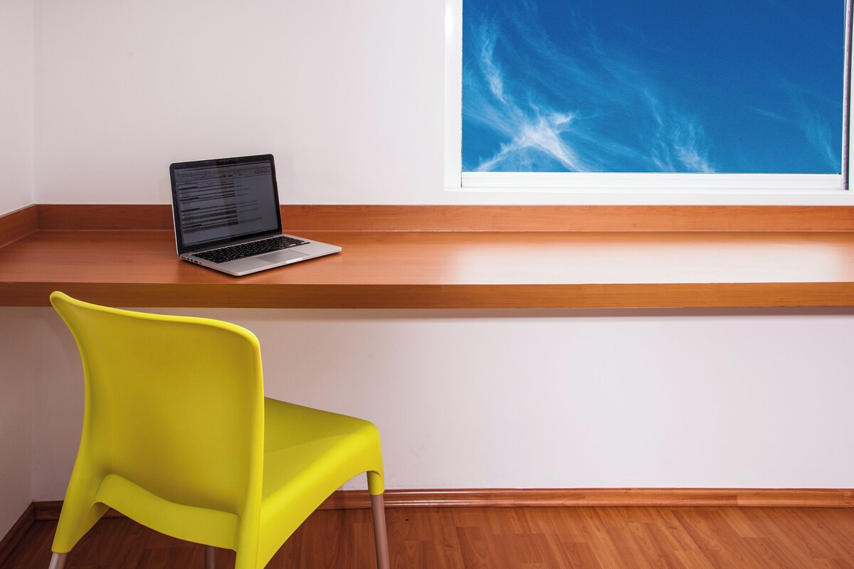 Una laptop sobre un repisa de madera, una silla de color amarillo brillante y una ventana con vista a un cielo azul.