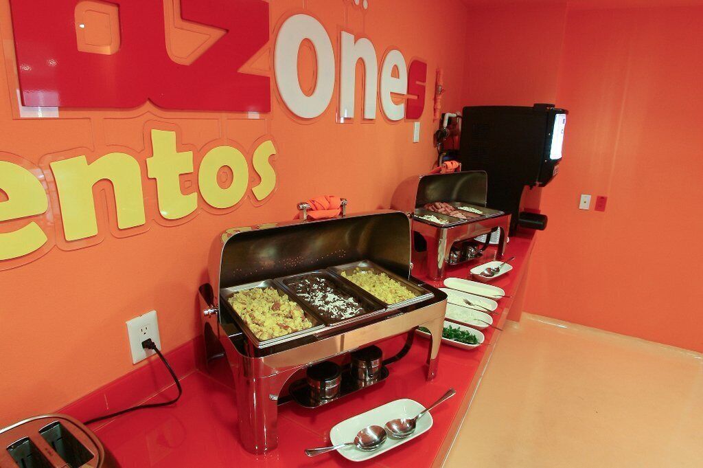 Zona de bufé con varias estaciones de comida, un televisor y una pared roja con señalización.