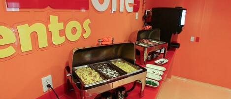 Zona de bufé con varias estaciones de comida, un televisor y una pared roja con señalización.