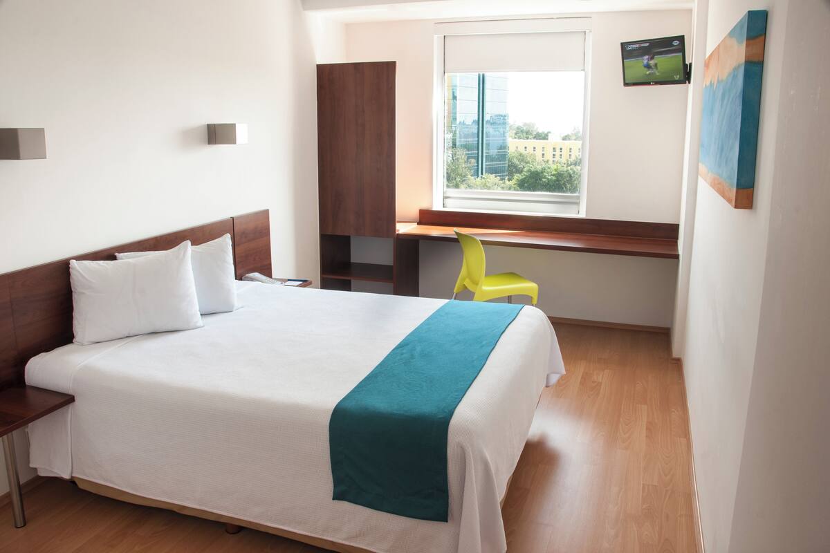 Una habitación de hotel con cama, escritorio, silla y televisor en la pared.