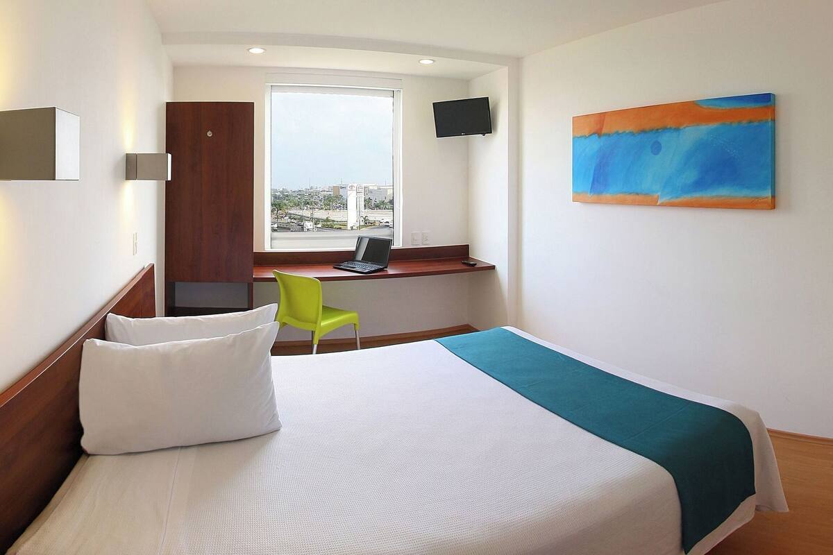 Habitación de hotel con una cama, un escritorio con una computadora portátil, una silla, un televisor y un cuadro en la pared.