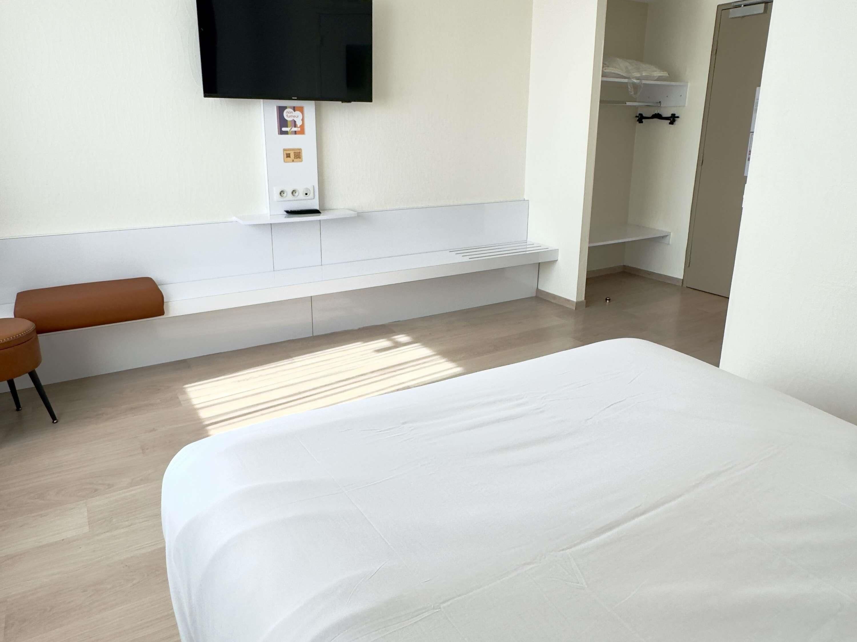 A hotel room with a bed, a bench, a television mounted on the wall, and a small shelf with a power outlet.