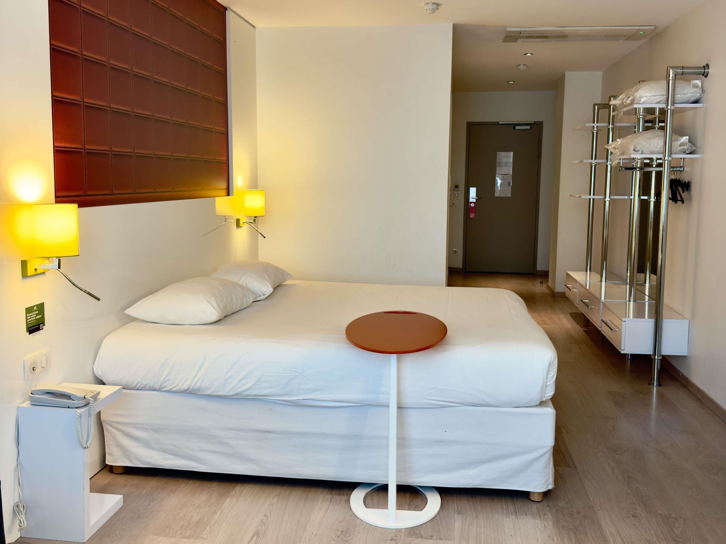 Une chambre d’hôtel comprenant un lit, des lampes de chevet, une petite table, une armoire et une porte donnant sur une autre pièce.