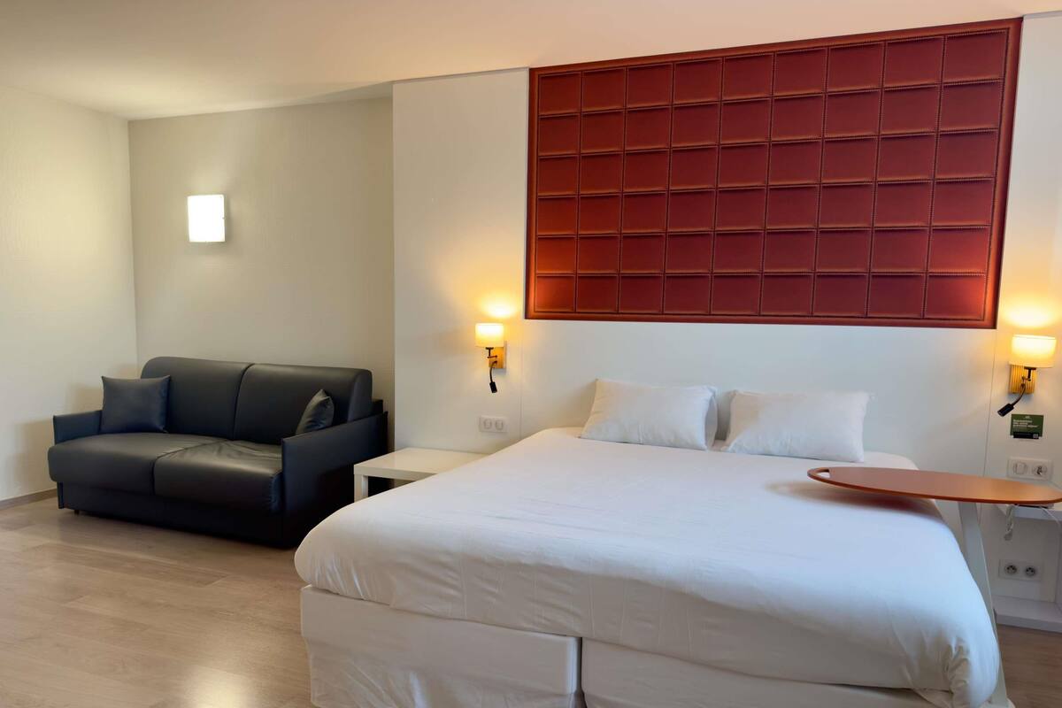 Una habitación de hotel con una cama grande, un sofá oscuro, dos mesitas de noche y un cabecero rojo.
