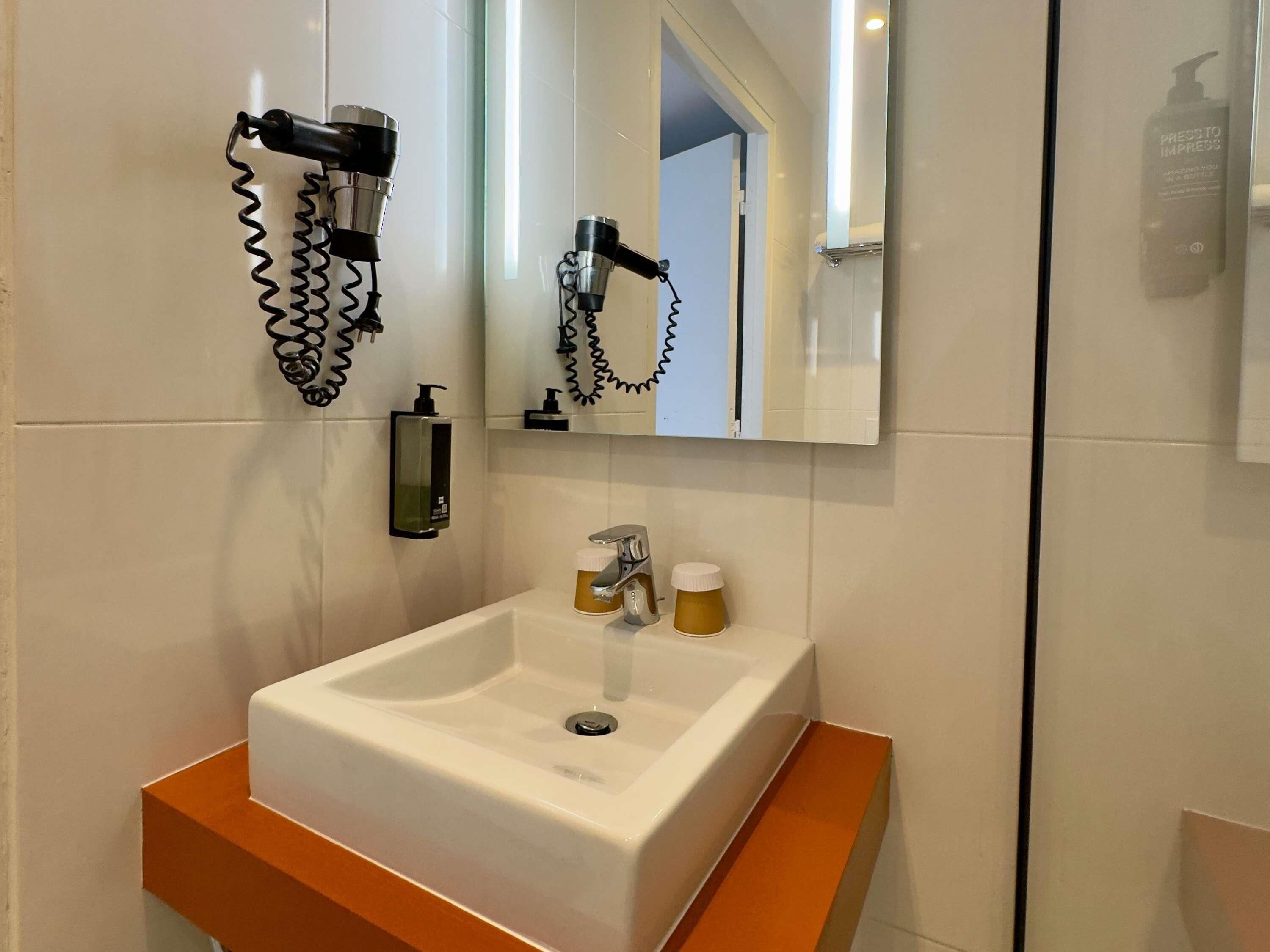 Un baño con lavamanos blanco, espejo, secador de pelo y dispensador de jabón.