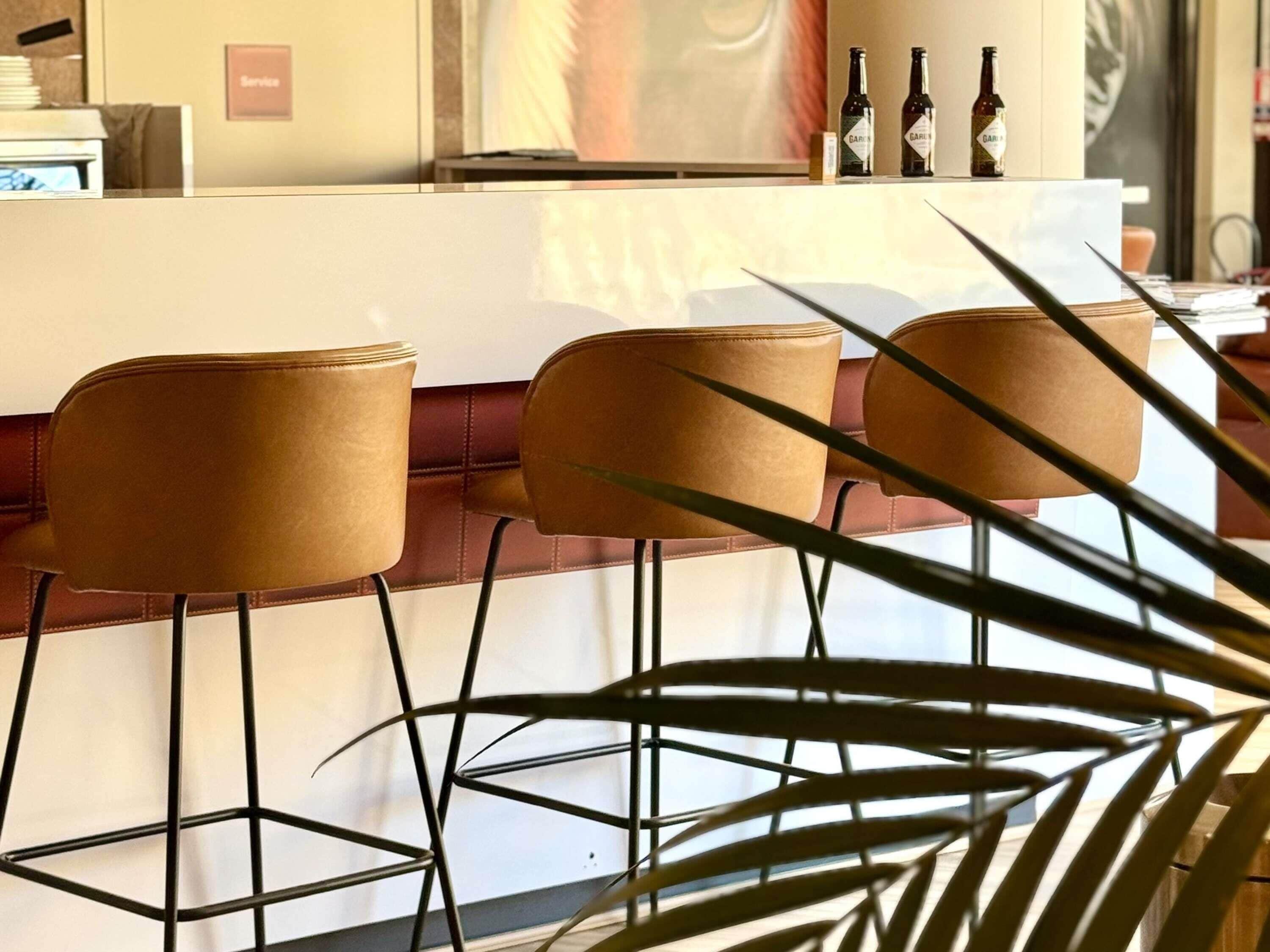 Una zona de bar con tres taburetes de cuero marrón, una barra metálica y botellas sobre la barra.