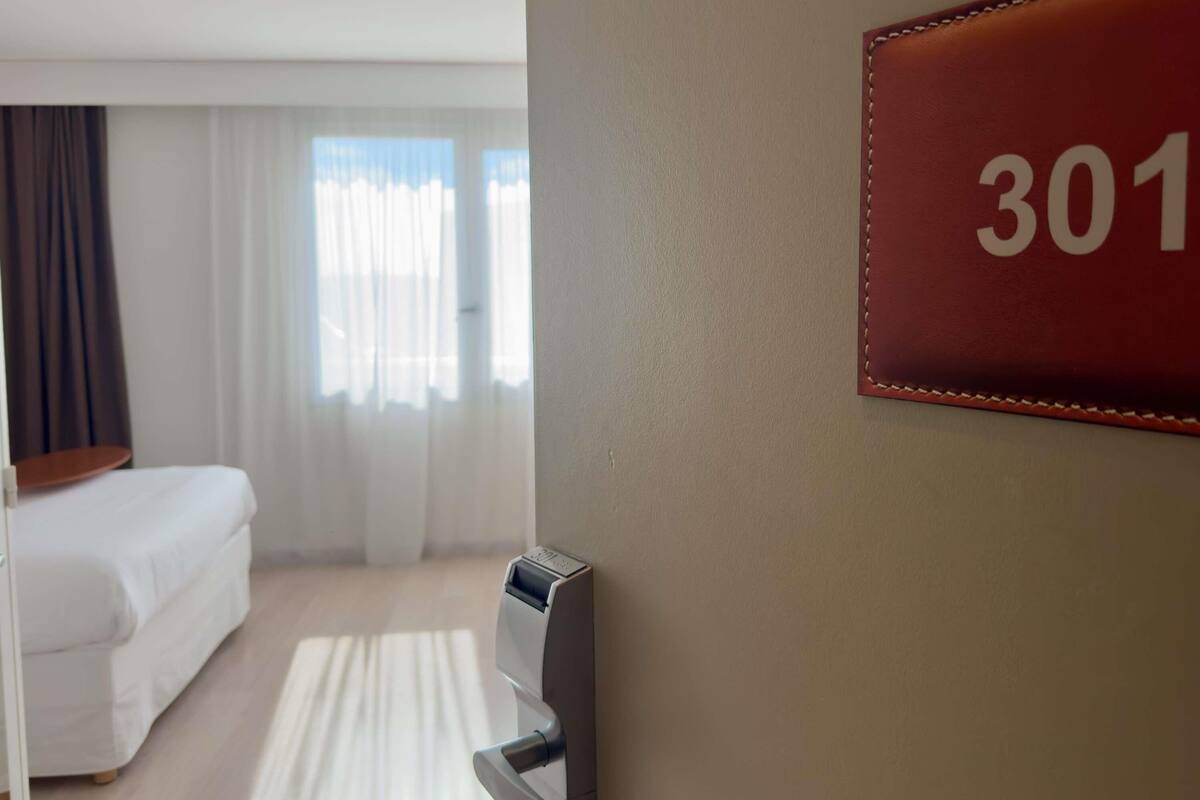 Habitación de hotel con cama, escritorio, un letrero con número en la pared y ventana con cortinas.