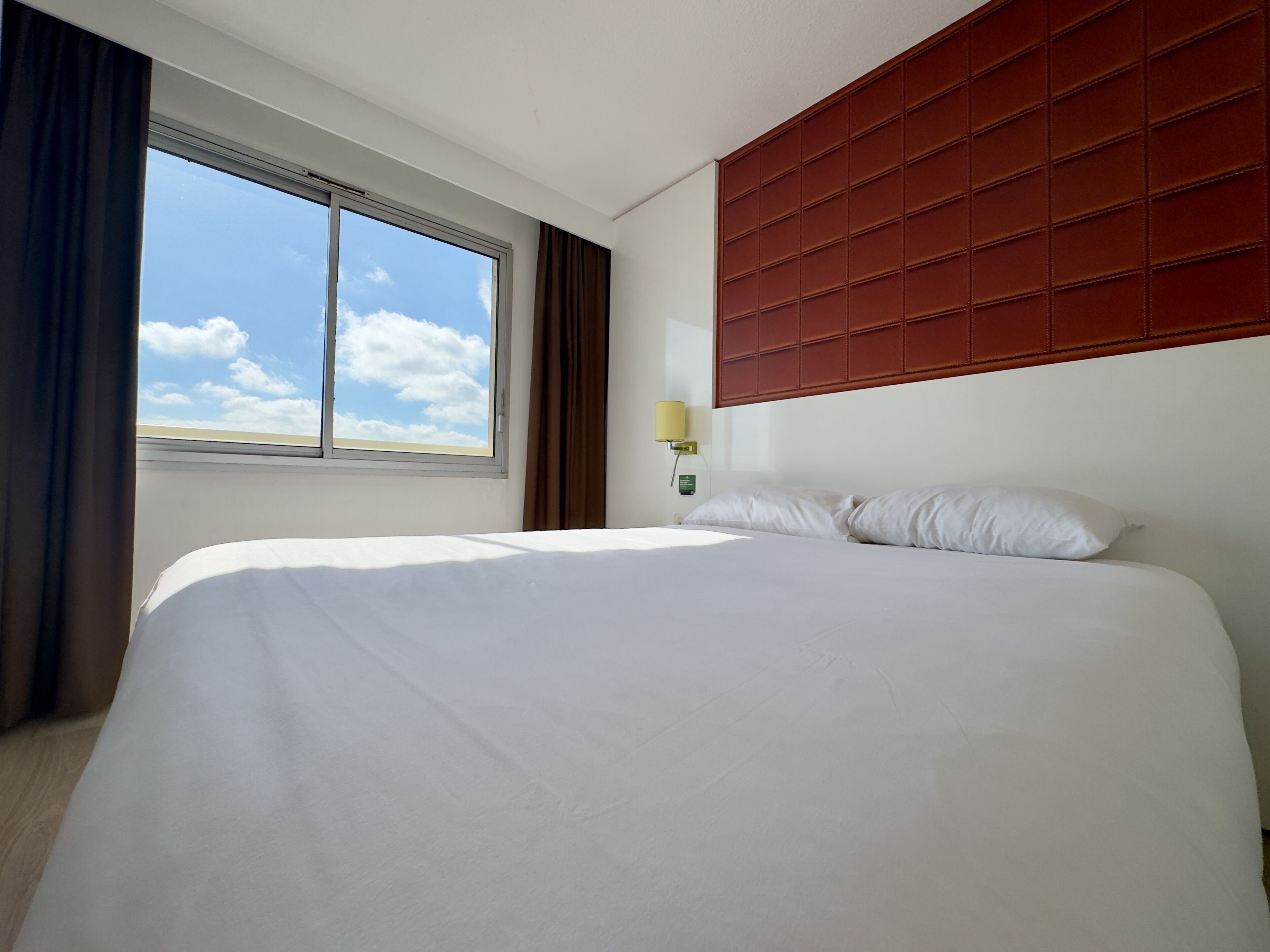 Hotelkamer met een groot bed, een raam met gordijnen, een wandlamp en uitzicht op de lucht.