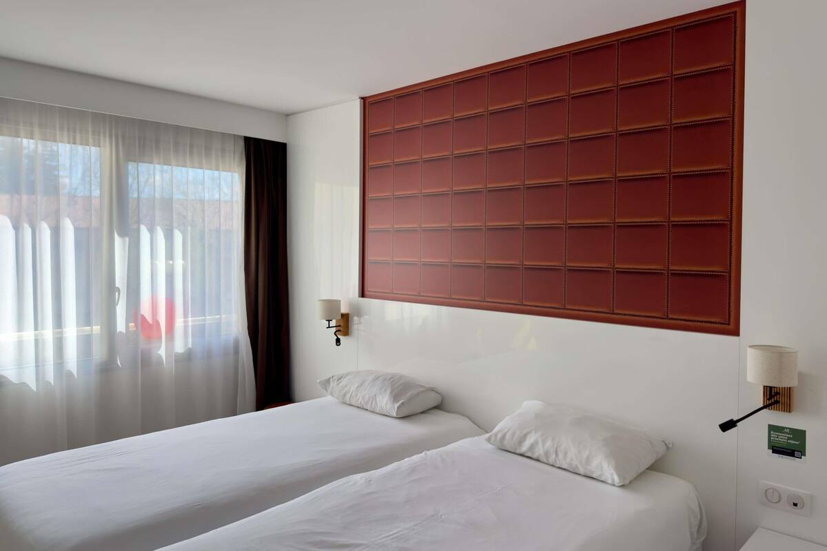 Habitación de hotel con dos camas, un ventanal con cortinas transl úcidas, cabecero rojo y lámparas fijadas a la pared.