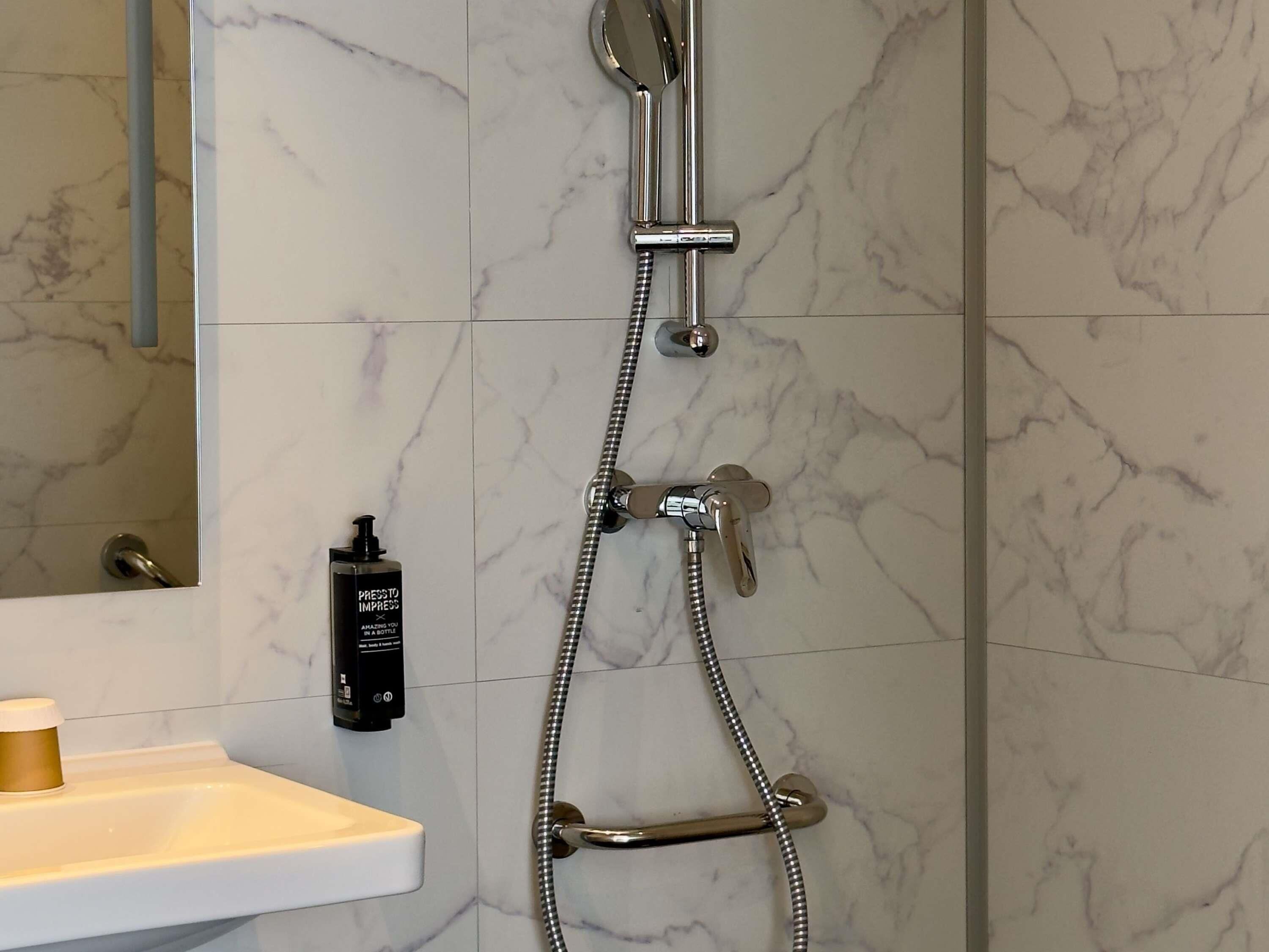 Une salle de bain avec un mur en marbre, une douche équipée d’un pommeau de douche à main et d’un flexible de douche, ainsi qu’une bouteille de shampoing fixée au mur.