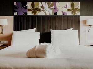 A hotel room with a bed, a headboard, a wall with floral artwork, and a nightstand with a lamp.