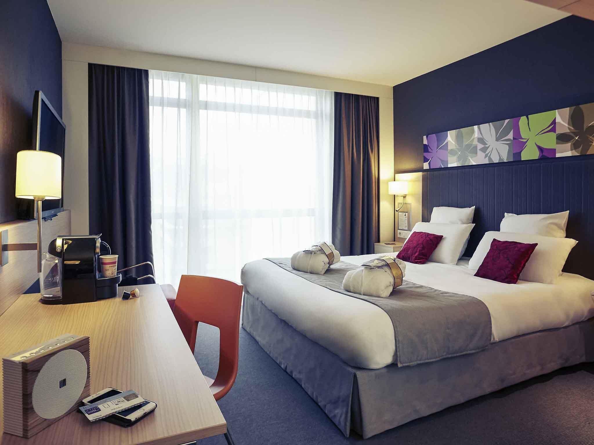 A hotel room with a bed, desk, chair, coffee maker, and a large window with curtains.