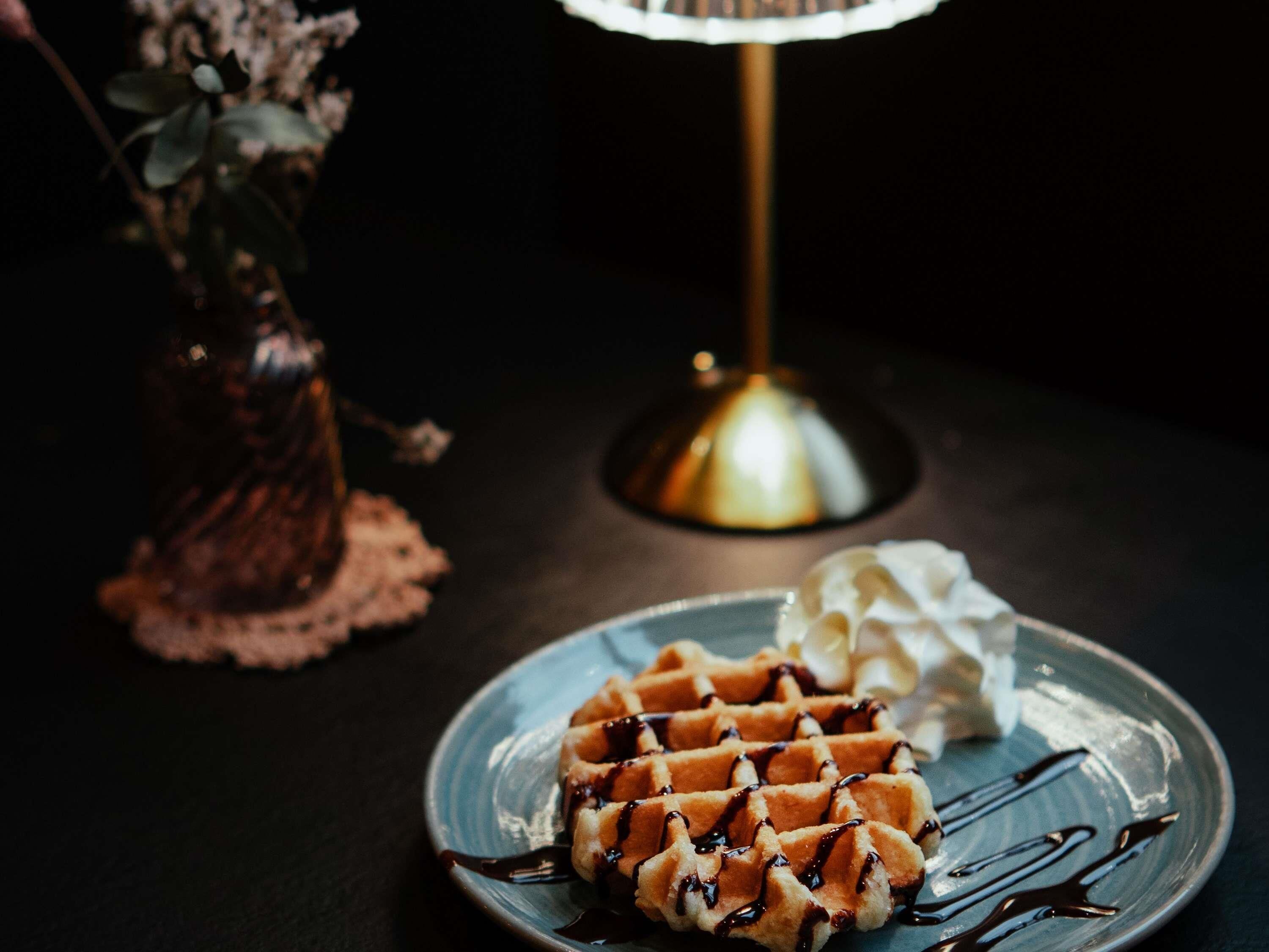 A waffle with chocolate syrup on a plate, a small vase with flowers, and a lamp on a table.