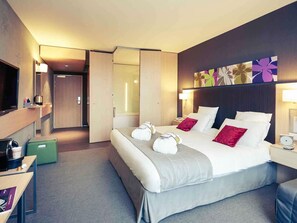 Habitación de hotel con cama, mesitas de noche, televisor, escritorio, una maleta verde y una puerta que da a un baño.
