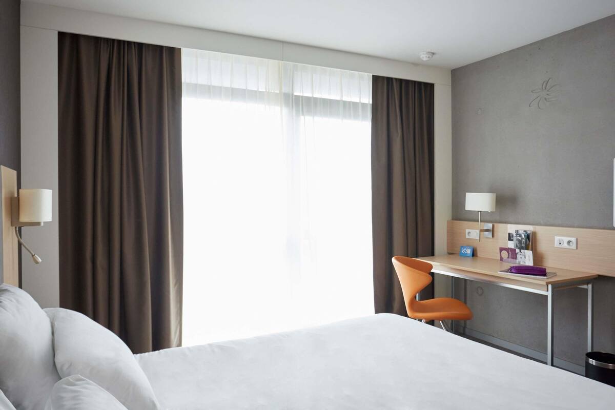 Une chambre d’hôtel avec un grand lit, un bureau avec une chaise, une petite table avec une lampe et une fenêtre avec des rideaux.
