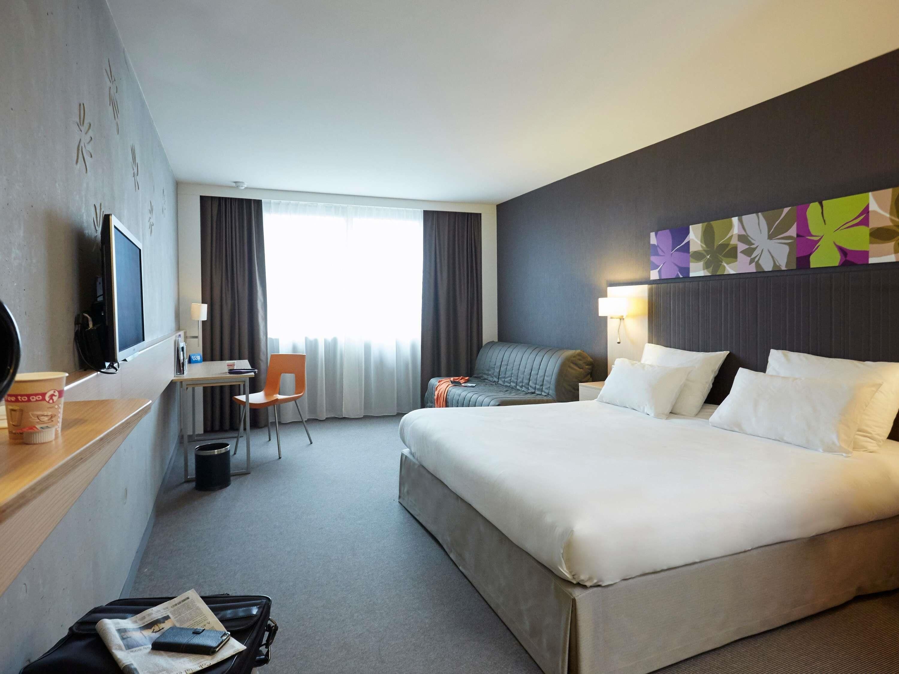 Une chambre d’hôtel avec un grand lit, un bureau avec une chaise, une télévision et une fenêtre avec des rideaux.