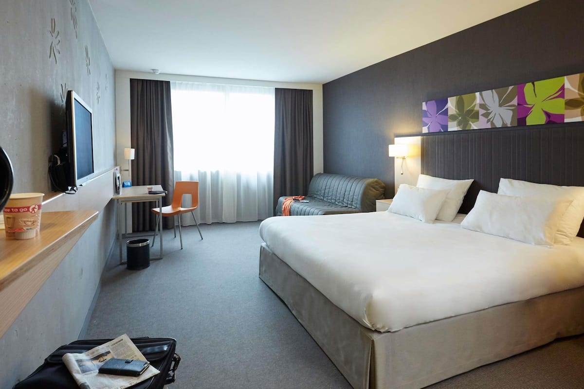 Une chambre d’hôtel avec un grand lit, un bureau avec une chaise, une télévision et une fenêtre avec des rideaux.