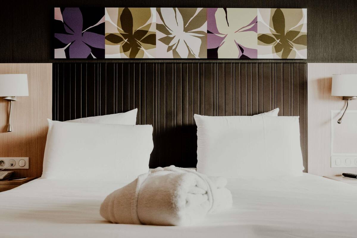 A hotel room with a bed, pillows, a towel, a nightstand, a lamp, and a wall with a floral pattern.