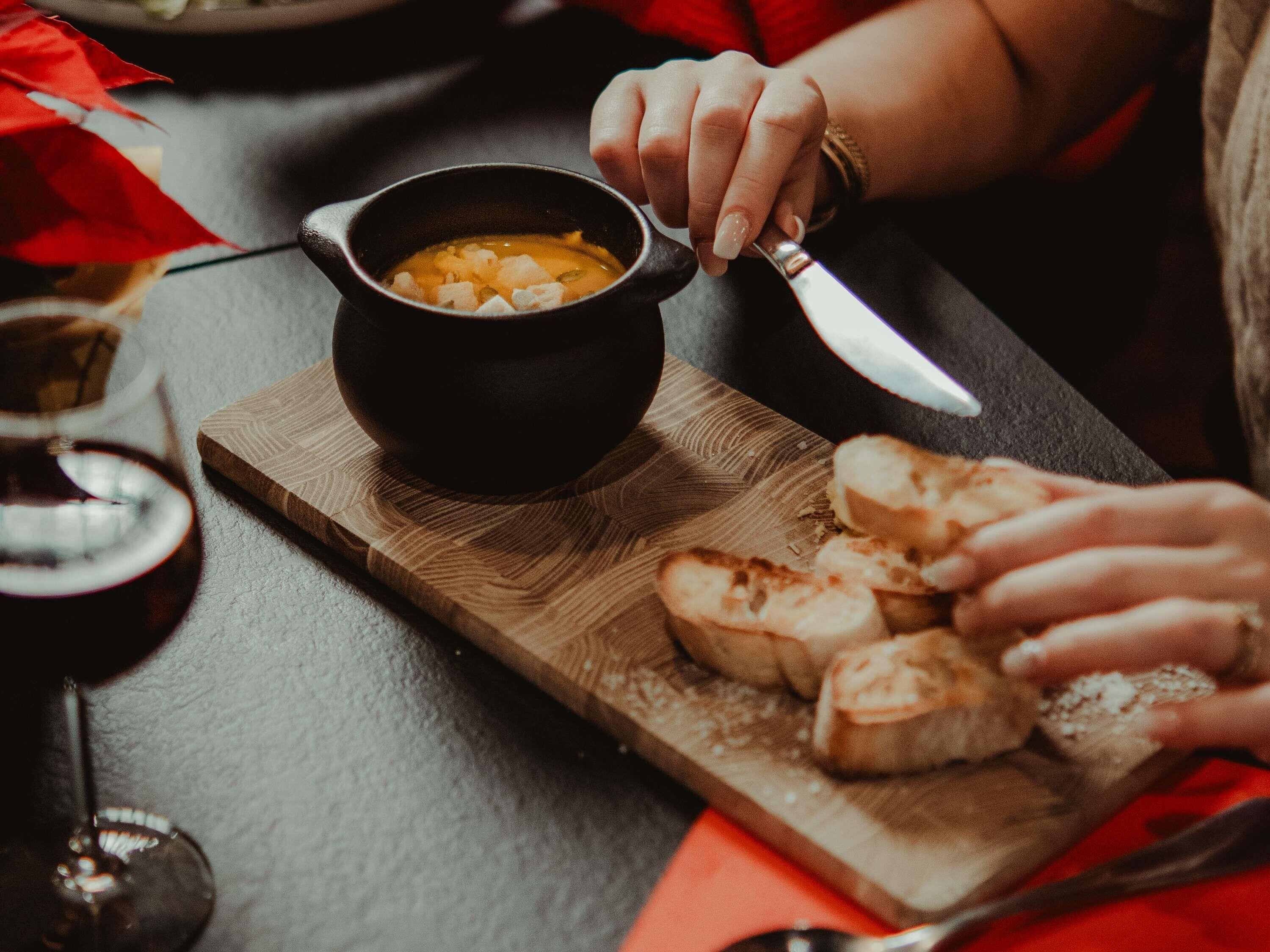 Una persona sosteniendo una cuchara sobre un tazón de sopa sobre una tabla de madera, con rebanadas de pan y una copa de vino tinto en la mesa.