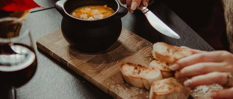 Una persona sosteniendo una cuchara sobre un tazón de sopa sobre una tabla de madera, con rebanadas de pan y una copa de vino tinto en la mesa.