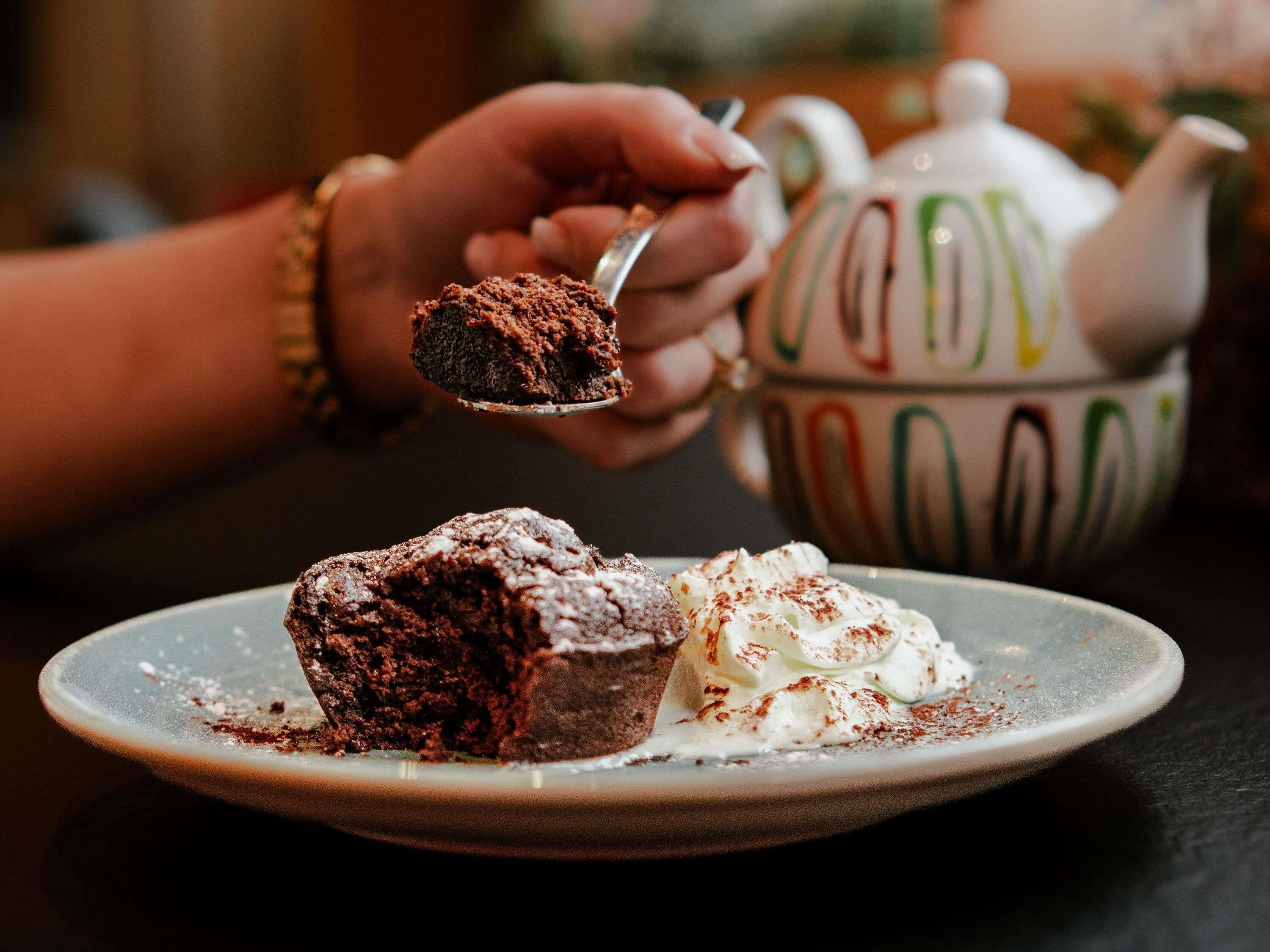 A dessert plate with a piece of cake and whipped cream, a teapot, and a person's hand holding a spoon.