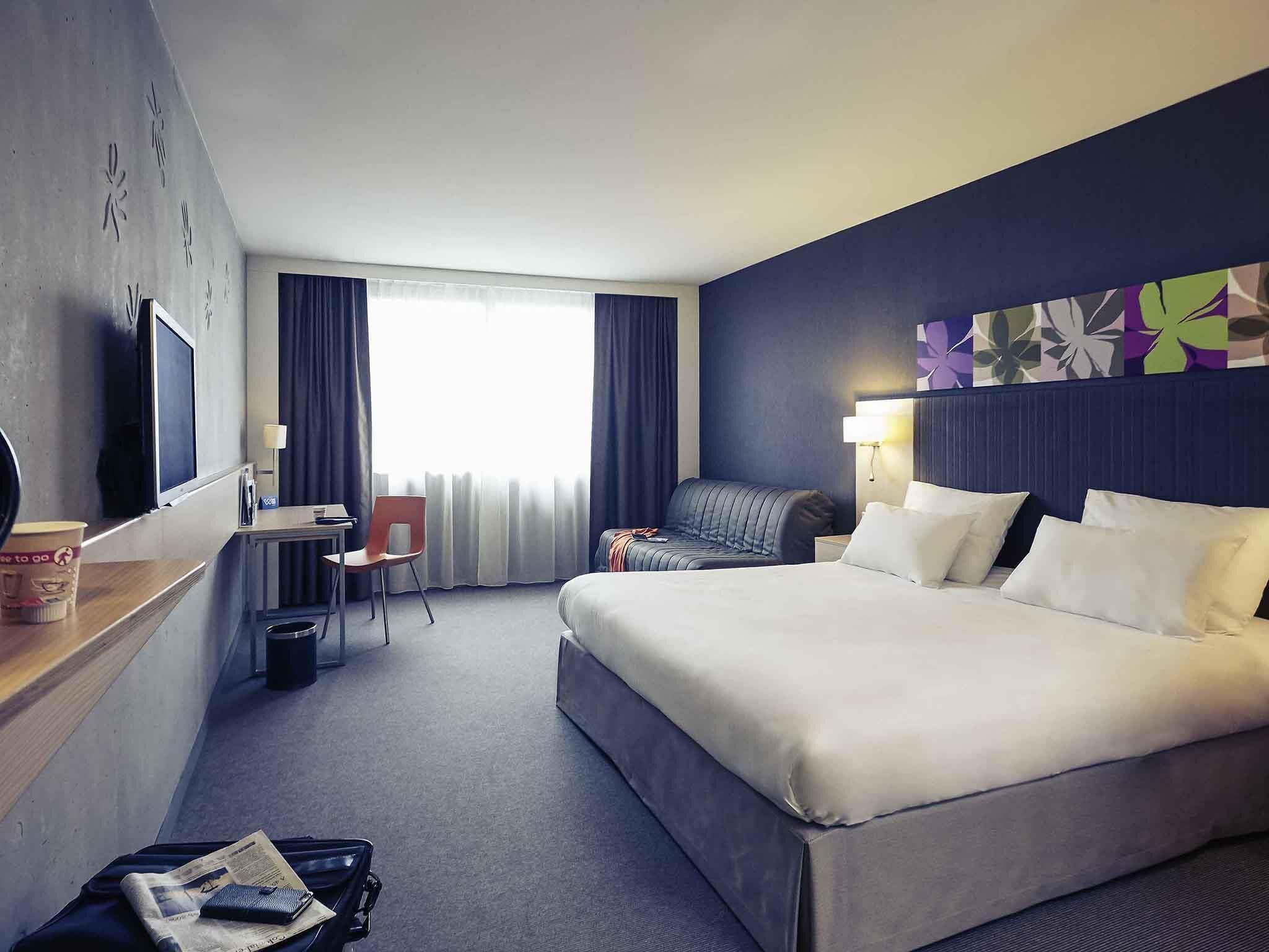 Ein Hotelzimmer mit einem großen Bett, einem Schreibtisch mit Stuhl, einem Fernseher und einem Fenster mit Vorhängen.