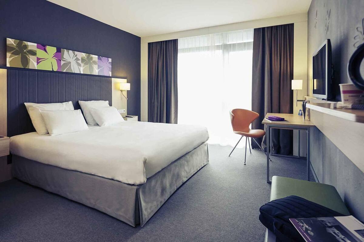 A hotel room with a bed, a desk, a chair, a TV, and a window with curtains.