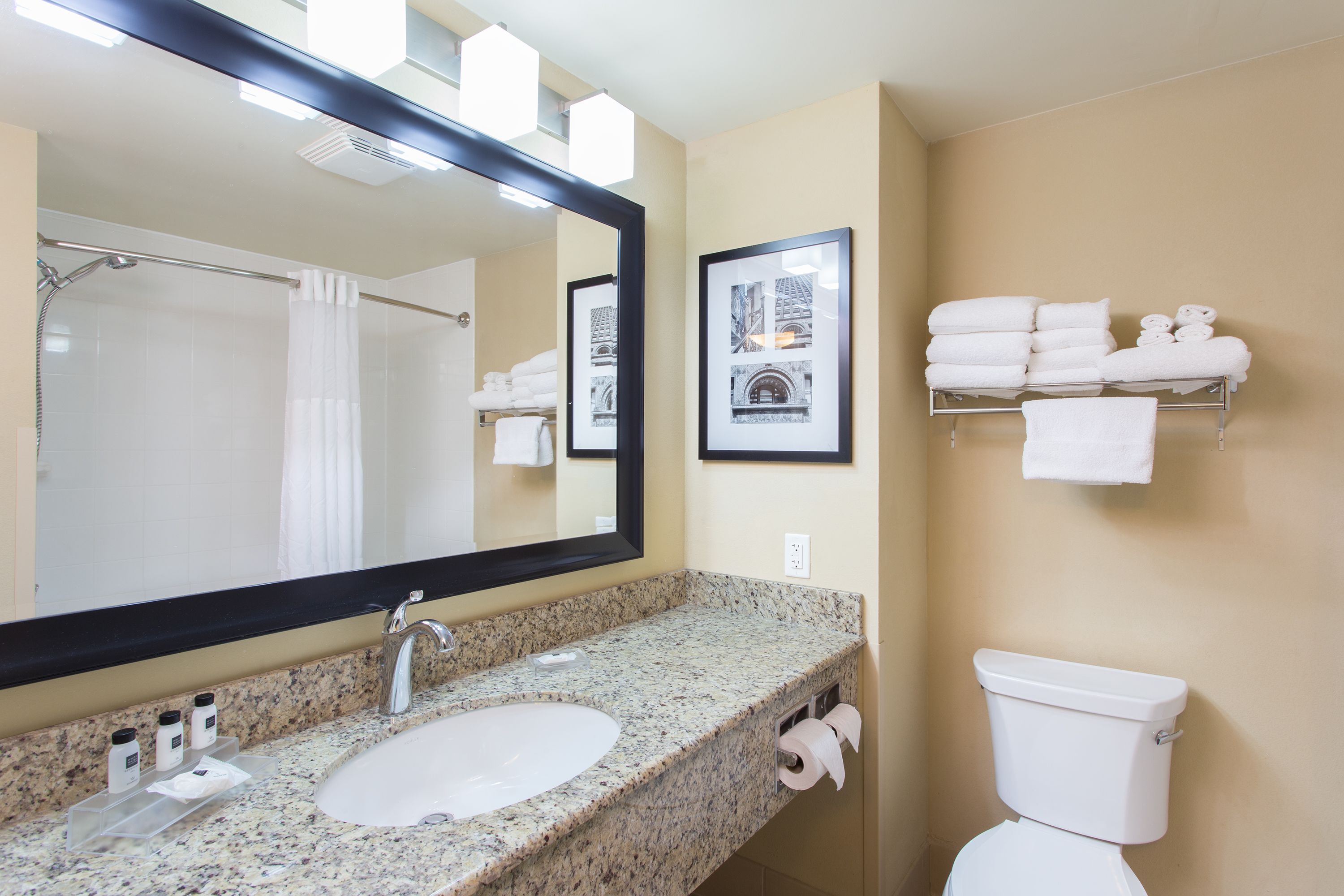 A bathroom with a granite countertop sink, a large mirror, a white toilet, and a towel rack with folded towels.