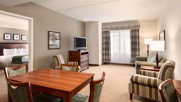 A hotel room with a wooden dining table, four upholstered chairs, a television, a sofa, a lamp, and a bed with a green throw.