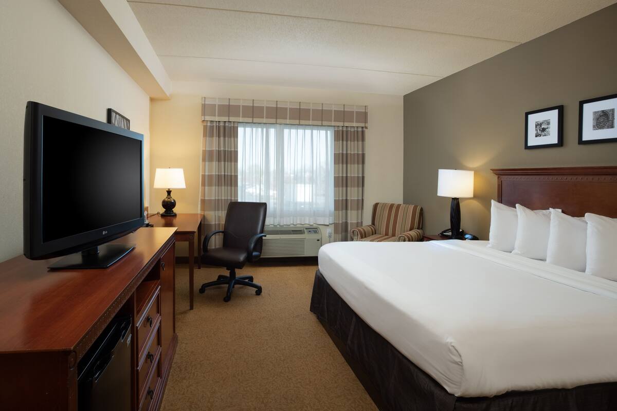 A hotel room with a large flat-screen TV, a bed with white linens, a desk with a lamp, a chair, and a window with curtains.