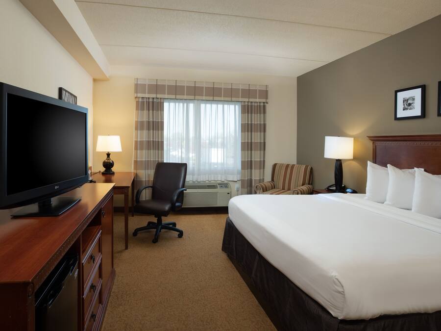 A hotel room with a large flat-screen TV, a bed with white linens, a desk with a lamp, a chair, and a window with curtains.
