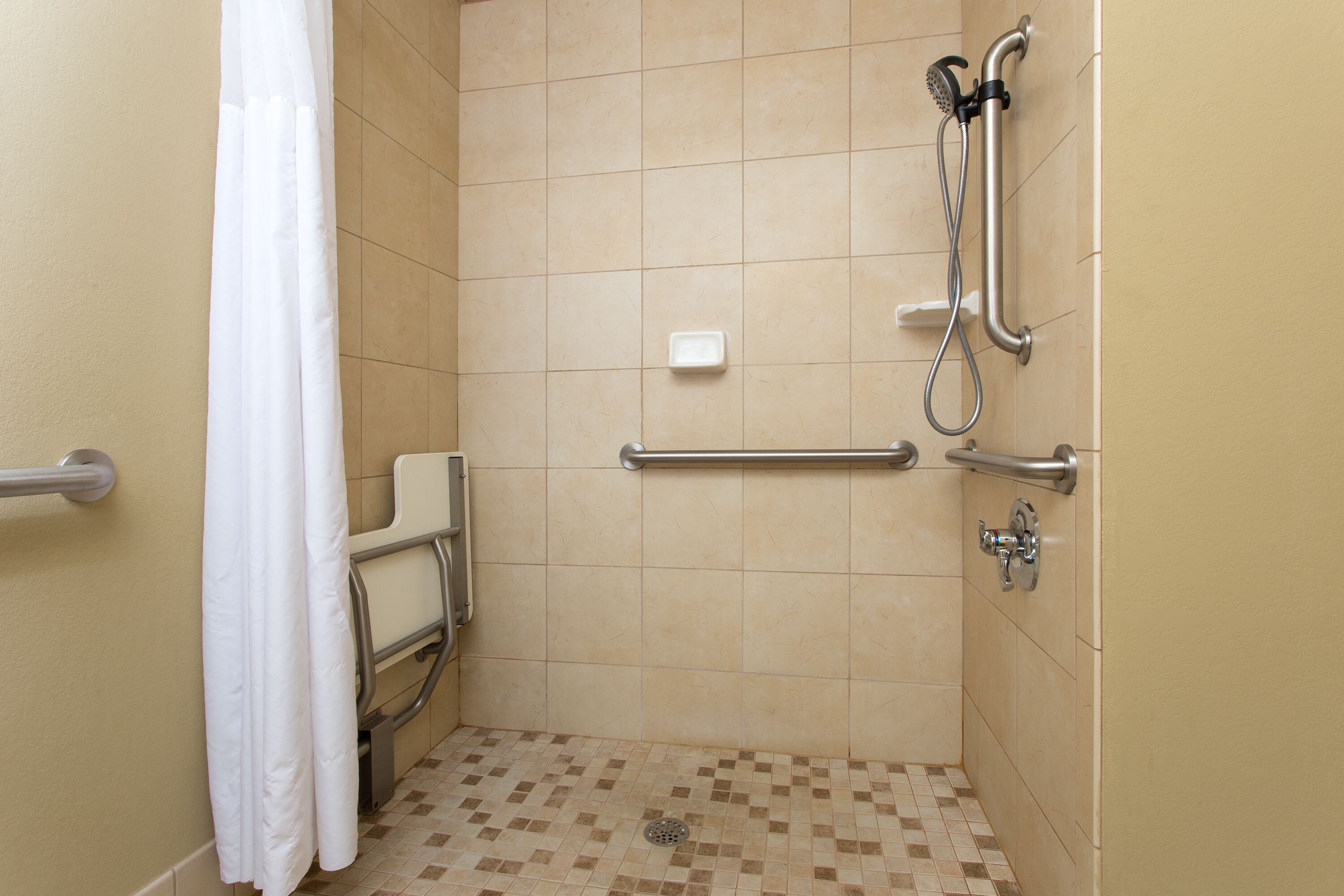 Een kleine, toegankelijke douche met een greep, een handdouche en een wit gordijn.