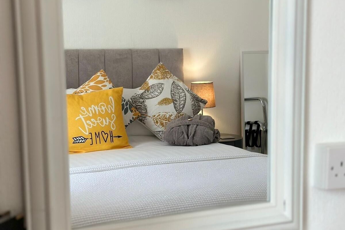 A bedroom with a bed, pillows, a bedside lamp, a mirror, and a bedside table with a tissue box.