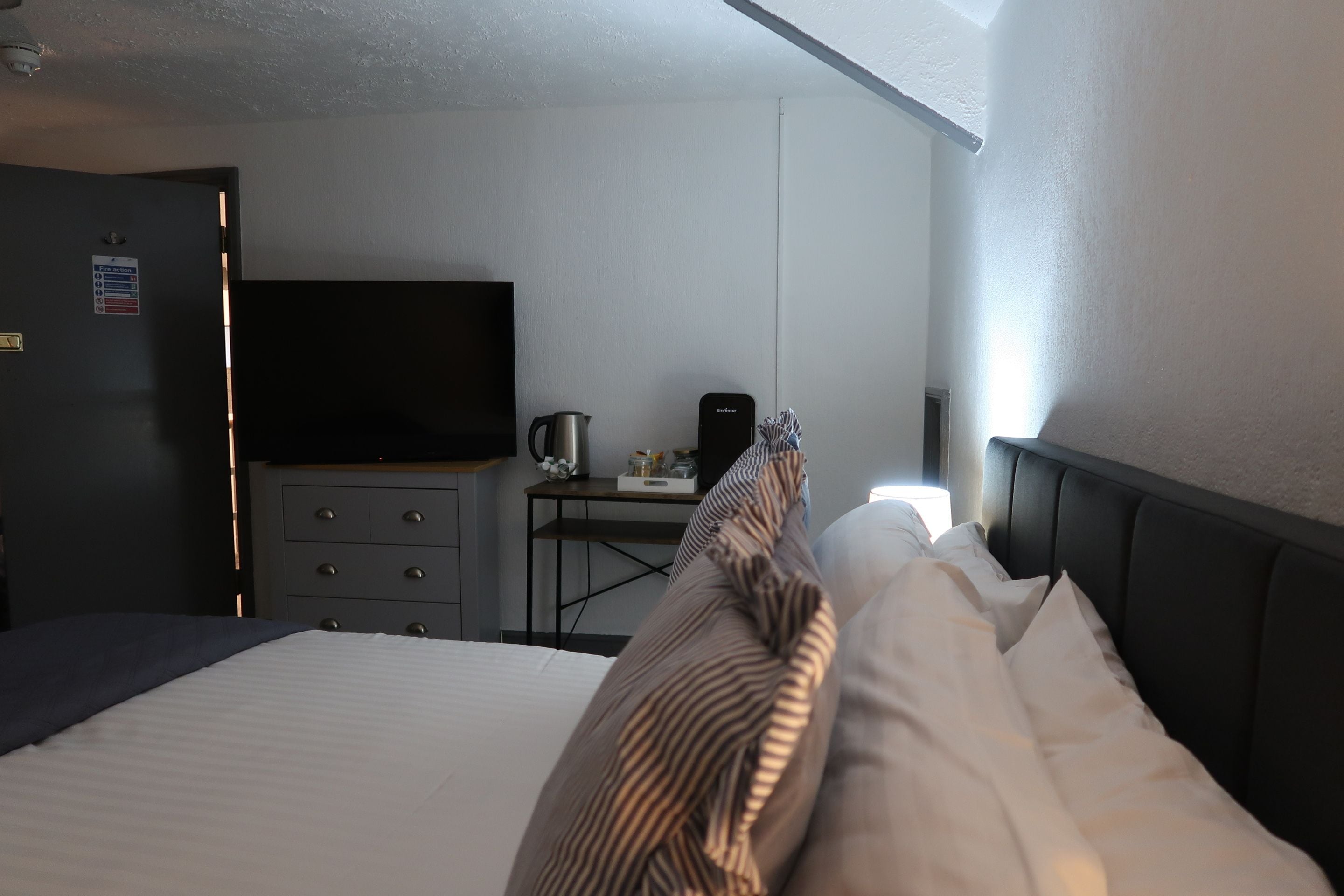 A hotel room with a bed, a desk with a kettle, a television, and a fire action sign on the door.