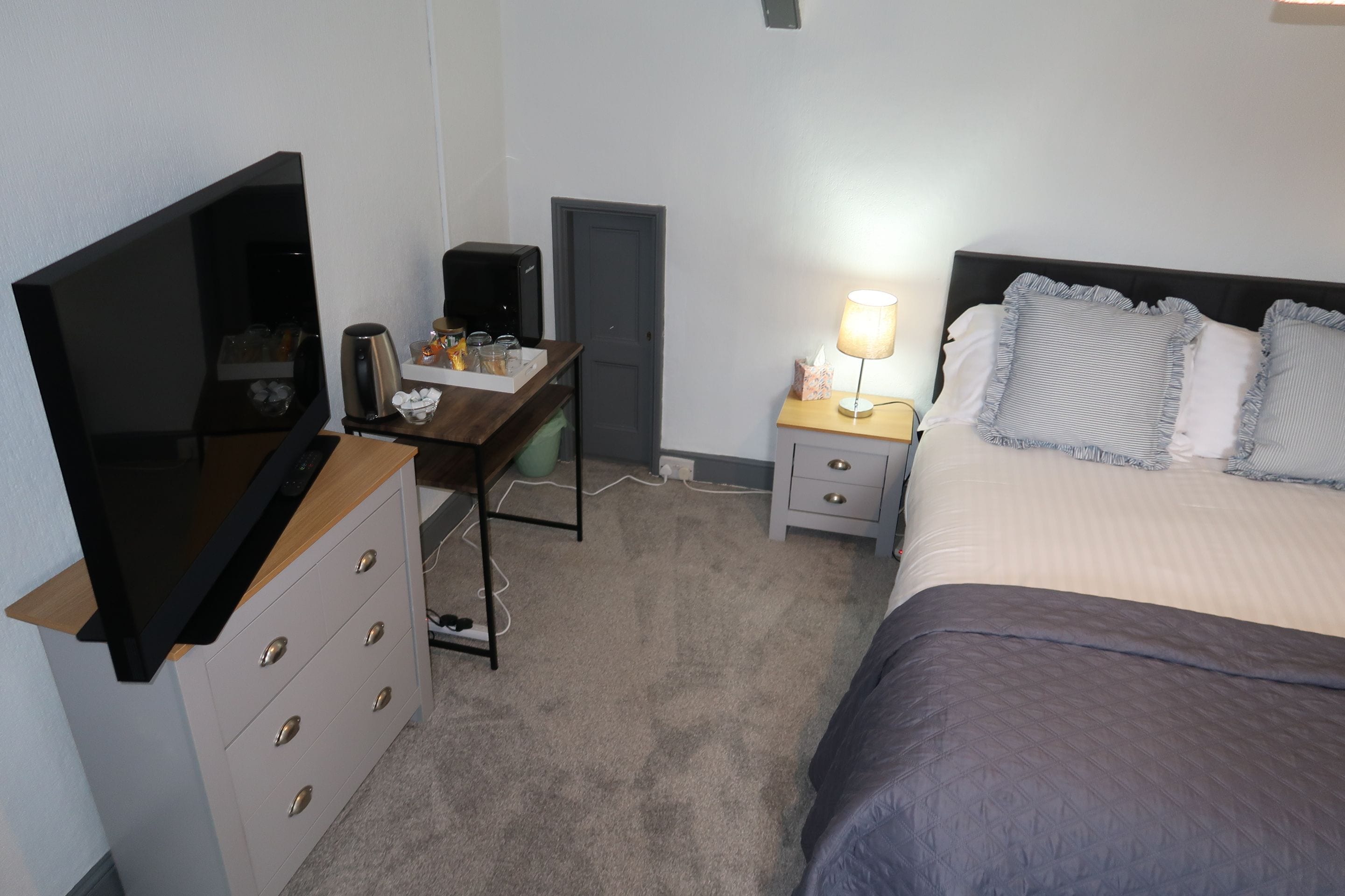 A hotel room with a bed, a desk with a computer, a TV, and a bedside table with a lamp.