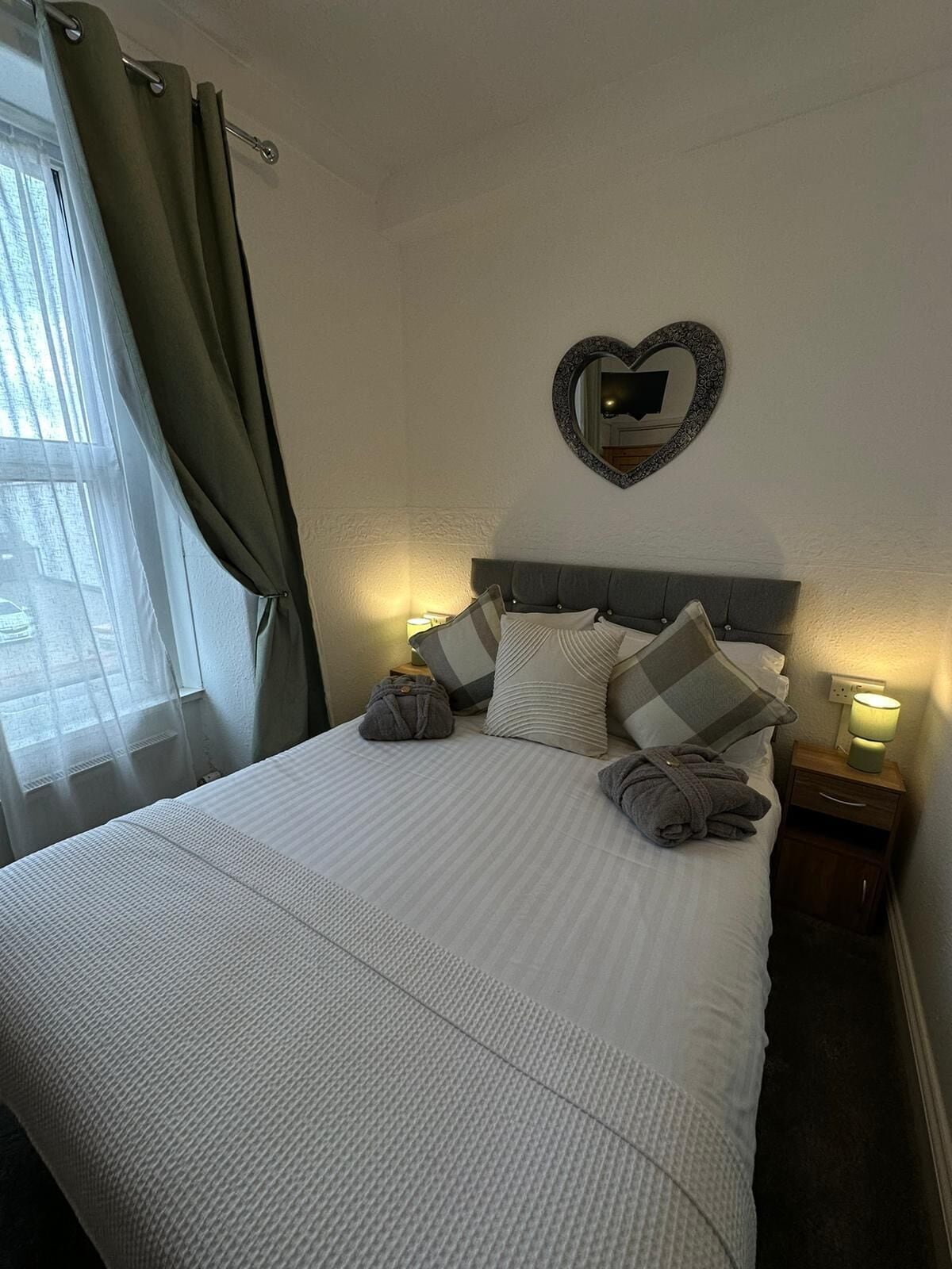 Un lit bien fait, avec un miroir en forme de cœur au-dessus, une lampe de chevet et une fenêtre avec des rideaux.
