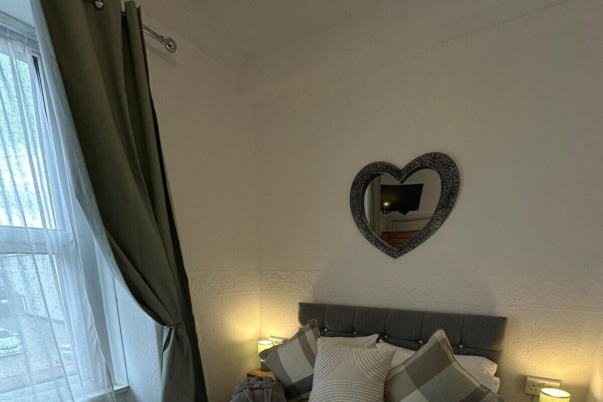 A neatly made bed with a heart-shaped mirror above it, a bedside lamp, and a window with curtains.