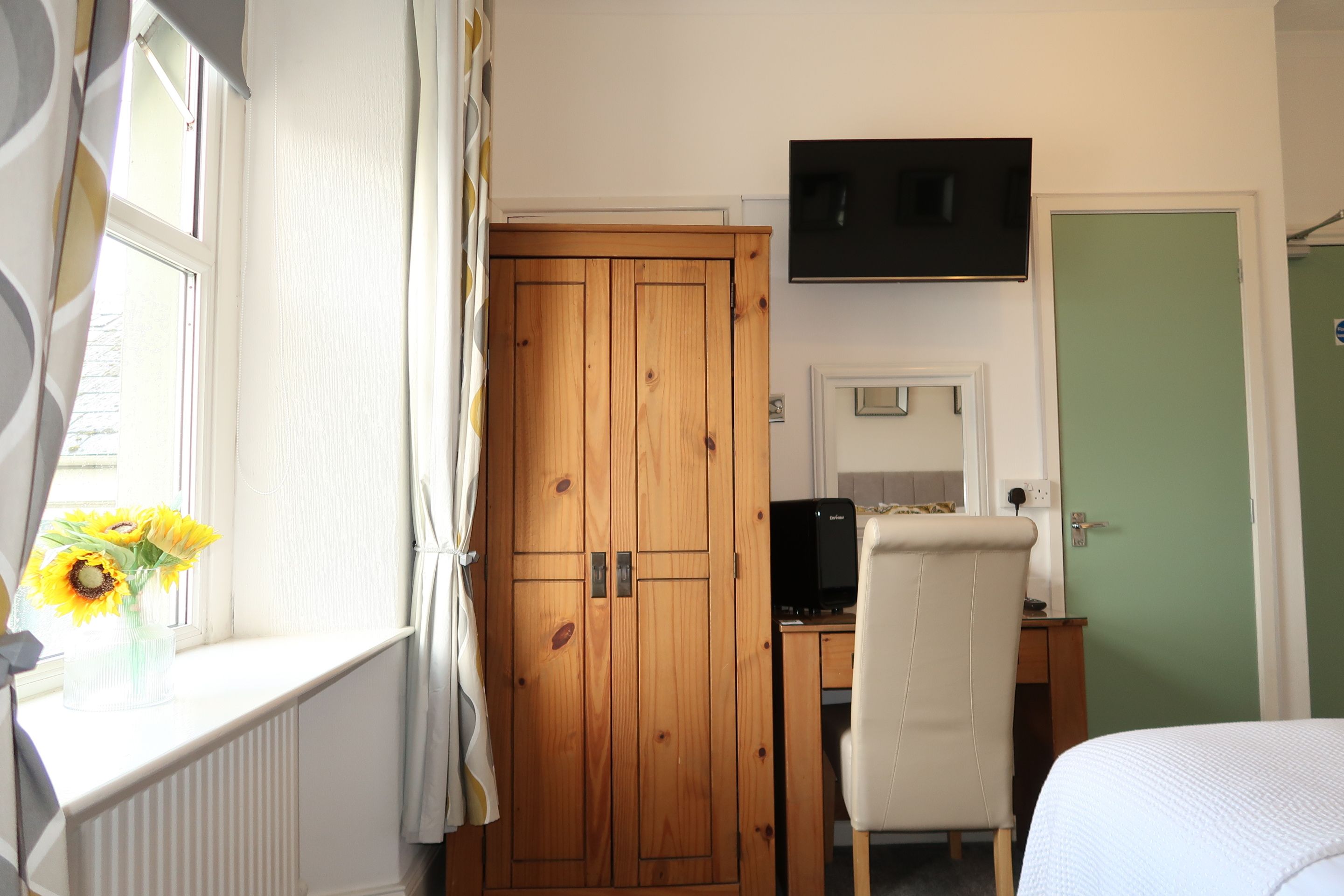 A room with a wooden wardrobe, a desk with a computer, a chair, a bed, and a window with curtains.